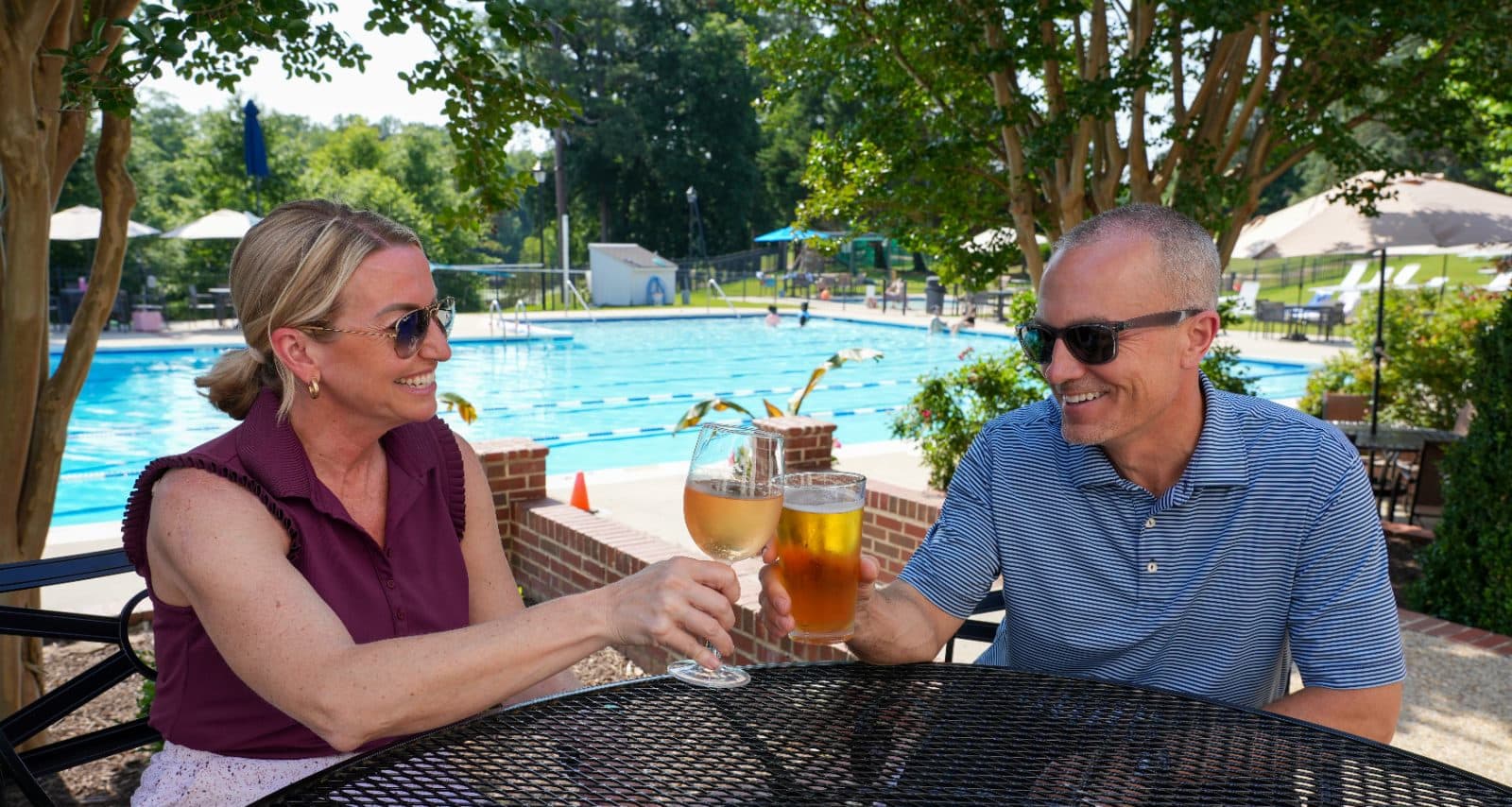 Two members raising a glass at a shaded table beside the pool